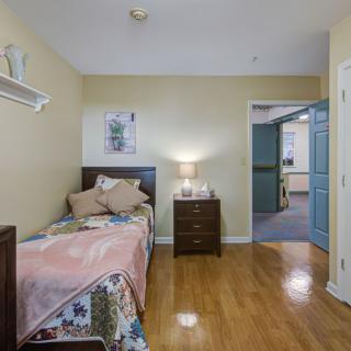 A cozy bedroom featuring a single bed with a colorful quilt, a nightstand with a lamp, and hardwood flooring.