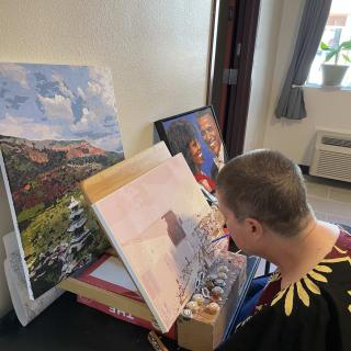 An artist working on a painting in their studio, surrounded by finished artworks.