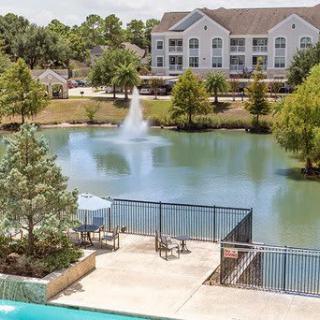 A serene outdoor setting featuring a lake with a fountain, surrounded by trees and buildings, with a swimming pool in the foreground.