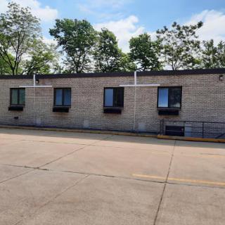 A view of a building with multiple windows, surrounded by trees and located next to a parking lot on a sunny day.