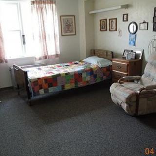 A cozy bedroom with a single bed covered by a colorful quilt, featuring a window with curtains, a recliner chair, and wall decorations.