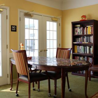 A cozy dining area with a round table, chairs, and a bookshelf, illuminated by natural light from large windows.