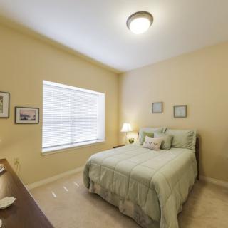 A cozy bedroom with a neatly made bed, soft lighting from a small lamp on the nightstand, and light streaming in through a window.