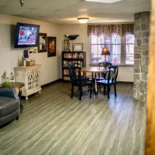A cozy living room featuring a small dining table, chairs, a wall-mounted television, and a bookshelf near the window.