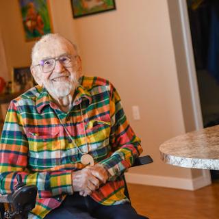 An elderly man with a warm smile sits in a colorful plaid shirt, enjoying a moment indoors.