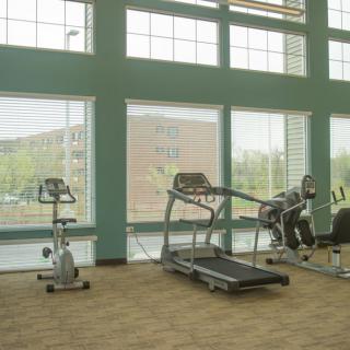 A gym room with various exercise equipment including a treadmill and a stationary bike, set against large windows.