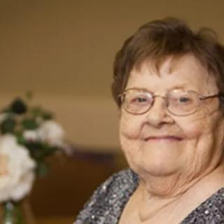 An elderly woman with glasses smiling indoors beside a flower arrangement.