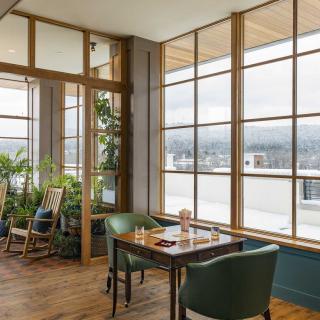 A cozy restaurant interior with large windows providing a scenic view, featuring wooden floors and lush indoor plants.