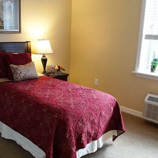 A cozy bedroom featuring a single bed with a red cover, a bedside lamp, and a window allowing natural light.