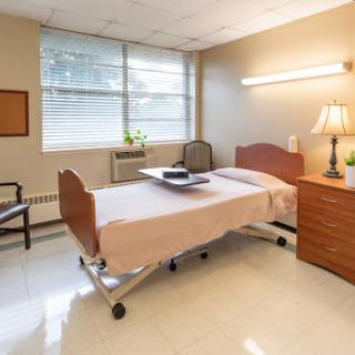 A well-lit hospital room featuring a single bed, a bedside table with a lamp, and a chair.