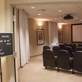 A small theater room with rows of chairs facing a large screen and projector.
