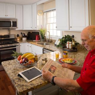 An elderly man reads a newspaper in a modern kitchen, surrounded by fresh fruit and technology.
