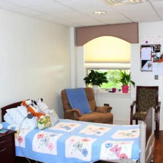 A cozy hospital room with a bed adorned with a floral blanket, an armchair, plants on the windowsill, and personal decor on the wall.