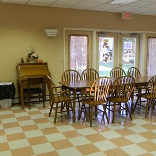 A dining area with a large table and multiple wooden chairs, featuring tiled flooring and windows in the background.