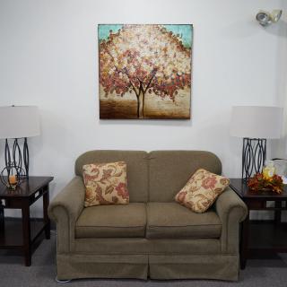 A cozy living room setup featuring a sofa with floral cushions, flanked by side tables and lamps, and adorned with a tree-themed painting on the wall.