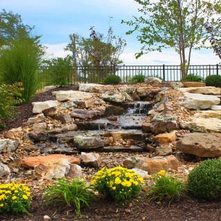 A landscaped garden featuring a small waterfall cascading over rocks, surrounded by lush greenery and vibrant flowers.