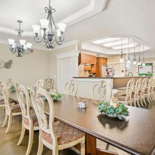 An elegant dining room with a long table and multiple chandeliers, overlooking a kitchen.