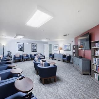 A spacious lounge area with comfortable seating, featuring individual chairs and small tables, a television, and bookshelves lining the walls.