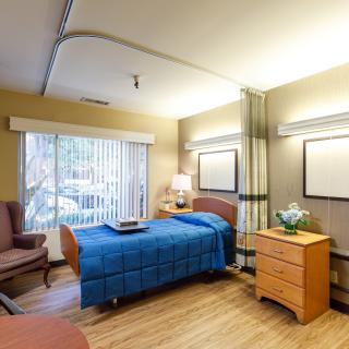 A hospital room with a single patient bed, wooden furniture, and a window with vertical blinds.