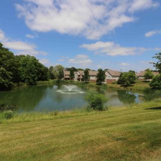 A scenic view of a pond with a fountain, surrounded by grassy slopes and apartment buildings under a clear blue sky.