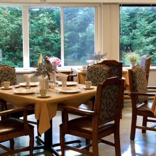 A cozy dining room with elegantly set tables and chairs, overlooking a lush green outdoor view through large windows.