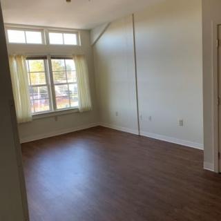 An empty room with wooden flooring and large windows allowing natural light.