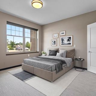 A modern bedroom with neutral tones featuring a bed, rug, and decorative wall art, illuminated by natural light from a large window.