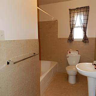 A simple bathroom with a bathtub, toilet, and pedestal sink, featuring tiled walls and a window with plaid curtains.