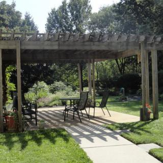 A shaded pergola over an outdoor patio with seating, surrounded by a lush garden on a sunny day.