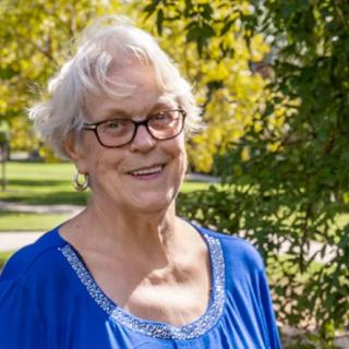 An elderly woman with glasses, smiling outdoors in a blue top surrounded by trees.