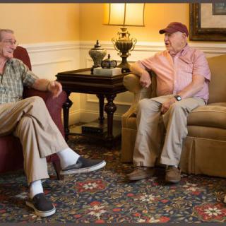 Two elderly men sit in a living room, engaged in conversation.