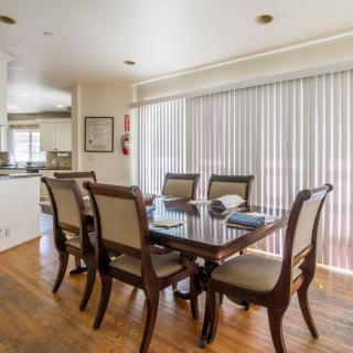 A modern dining room with a glass-topped table and six chairs, featuring sliding doors with vertical blinds and a hardwood floor.
