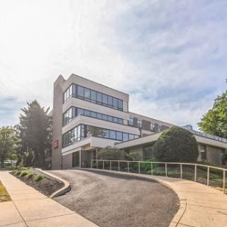 A modern building set amidst lush greenery, featuring large windows and a gently sloping access road.
