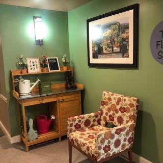 A cozy corner with green walls, featuring floral-patterned chair and wooden desk, complemented by artwork and decorative items.