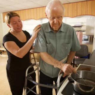 A caregiver assists an elderly man using a walker in the kitchen.