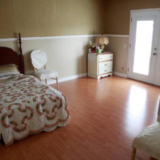 A minimalist bedroom with wooden flooring, featuring a bed, a chair, and a dresser with a lamp.