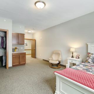 A cozy bedroom with a bed, chair, and lamp, featuring neutral colors and carpet flooring.