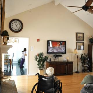 A cozy living room interior with a television mounted on the wall, a large clock, and a wheelchair facing the TV.