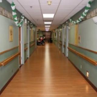 A long, well-lit hallway with wooden floors and green walls, typical of a hospital or nursing home.