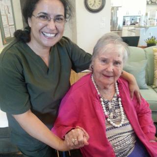 An elderly woman in a pink sweater sits in a wheelchair, holding hands with a smiling caretaker.