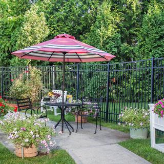 A cozy garden patio setup with a striped umbrella, table, and chairs surrounded by lush greenery and flowers.