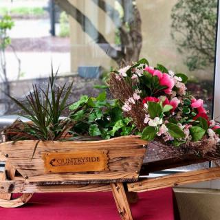 A decorative wooden wheelbarrow filled with colorful pink flowers and lush green plants, placed indoors.