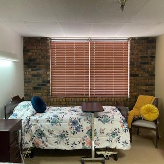 A serene hospital room featuring a neatly made bed with floral bedding, a cozy chair, and natural light filtering through wooden blinds.