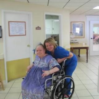 An elderly woman in a wheelchair smiling with a healthcare worker in a nursing home setting.