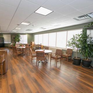 A spacious lounge area with tables and chairs, featuring a wooden floor and large windows allowing natural light to enter.