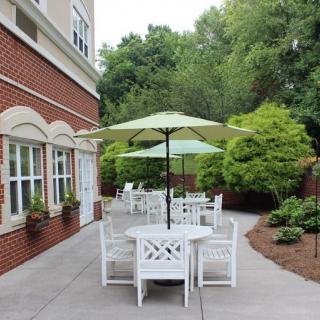 An outdoor patio area with white tables and chairs shaded by green umbrellas, surrounded by lush greenery next to a building.