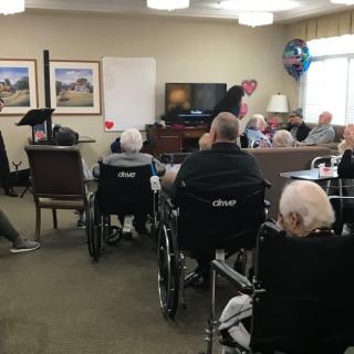 A group of elderly people gathered in a nursing home for a communal activity, some seated in wheelchairs.