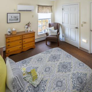 A cozy bedroom with a patterned bedspread, wooden dresser, and armchair by the window.
