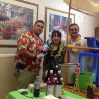 Three people dressed in tropical attire at a luau-themed party with drinks and decorations.