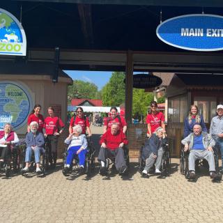 A group of people in wheelchairs with others standing behind them, gathered at an outdoor entrance.
